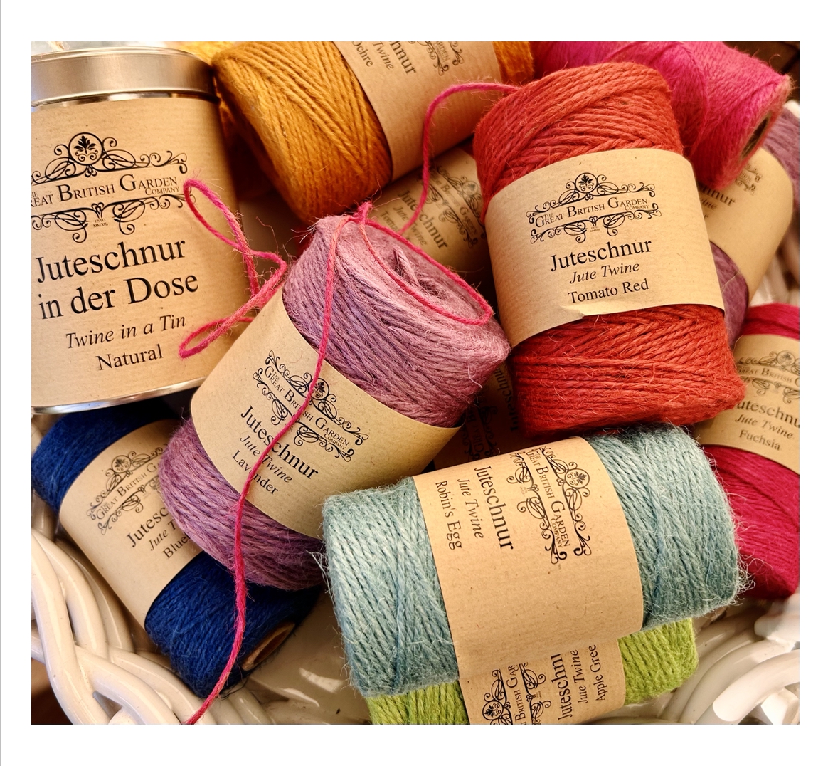 Bunte Juteschnüre: naturfarbene Schnur in Metalldose und viele kleine Spulen in Rot, Lila, Blau, Grün und Türkis. Jede Spule hat braunes Etikett mit Produktnamen und Logo, dekorativ im hellen Korb für Garten.