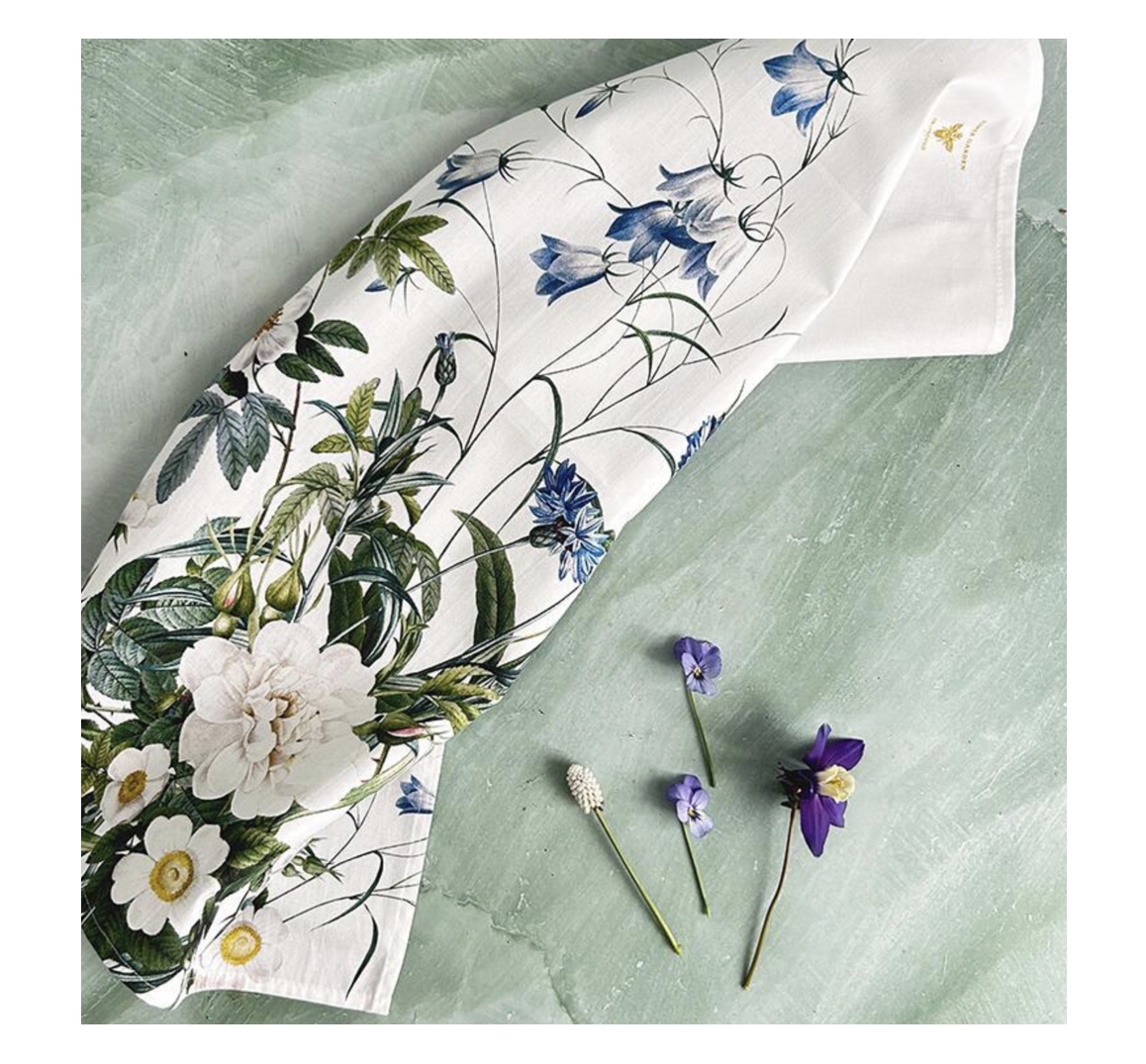 Gefaltetes weißes Bio-Baumwoll-Geschirrtuch mit üppigem BLUE FLOWER GARDEN Blumenmuster: blaue Glockenblumen, weiße Rosen und grüne Blätter. Liegt auf mintgrüner Oberfläche, daneben drei frische Blüten in Violett und Weiß.